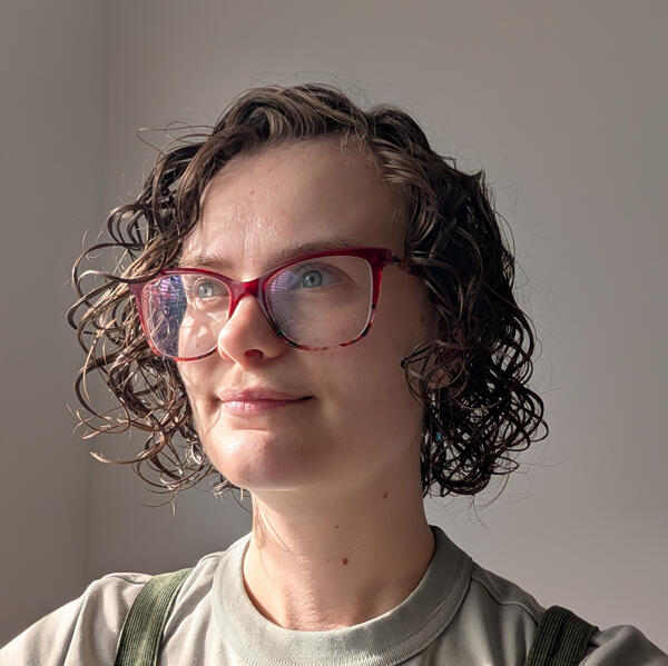 Kirsty Headshot Cropped headshot of Kirsty. They are a white person with chin-length curly brown hair, wearing a light green t-shirt and a pair of red glasses.
